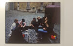Skan analogowego zdjęcia; grupa osób siedzi na brukowanej ulicy w okręgu; dyskutują ze sobą; jedna osoba robi notatki; w tle skromne zabudowania staromiejskie.