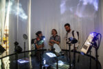 Grupa trzech osób podczas dyskusji przy obiekcie; ciemny blat stołu, przy którym stoją, odbija rzucane na niego obrazy, pokryty jest warstwą wody.